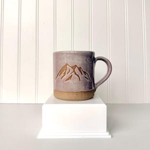 May include: A ceramic mug with a speckled lavender glaze on the upper portion and a textured, unglazed brown clay base. The front of the mug features a carved, brown mountain range graphic. It has a curved handle on the right side.