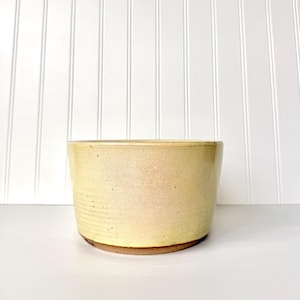 Pode incluir: Uma tigela cilíndrica de cerâmica com um esmalte amarelo pálido salpicado. O exterior apresenta uma secção superior lisa e uma série de estrias horizontais na base inferior. O rebordo é arredondado e a base mostra uma borda de argila castanha.