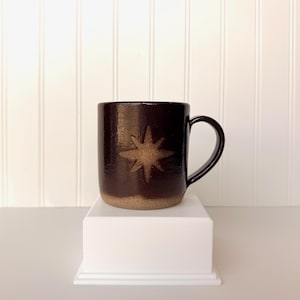 May include: A dark brown ceramic mug with a glossy finish and a textured handle. The front of the mug features a debossed eight-pointed star design with a speckled, light tan finish that matches the unglazed base of the mug.