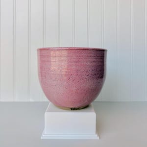 May include: A round ceramic bowl with a speckled pink glaze finish. The bowl features a smooth, curved silhouette with a slightly darker rim and a visible base. The exterior surface displays a textured, mottled appearance in various shades of pink.