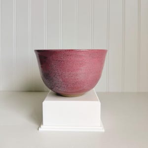 May include: A handmade ceramic bowl with a deep burgundy glaze. The bowl has a wide, rounded shape and sits on a white pedestal. The interior and exterior surfaces are a speckled, matte finish, perfect for serving food.