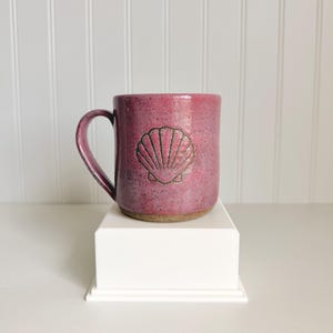 May include: A ceramic mug in a speckled magenta hue, featuring a carved seashell design. The mug has a rounded handle and sits on a white pedestal. The mug is a standard size for coffee or tea.