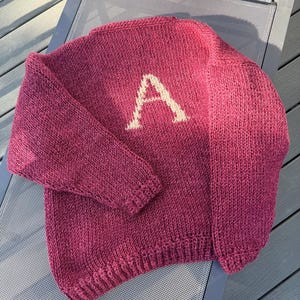 May include: A long-sleeved knitted sweater in a deep burgundy color featuring a prominent, light-colored letter A stitched into the center of the chest. The garment has a ribbed crew neckline, ribbed cuffs, and a ribbed hem, showcasing a textured knit.