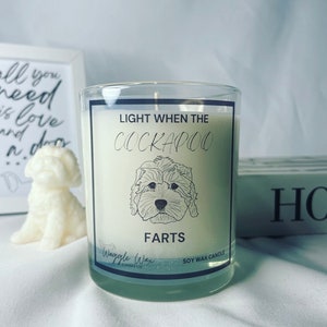 May include: A white soy wax candle with a black label that says "Light When The Cockapoo Farts". The label also features a black and white illustration of a cockapoo dog. The candle is made by Waggle Wax Candle Co.