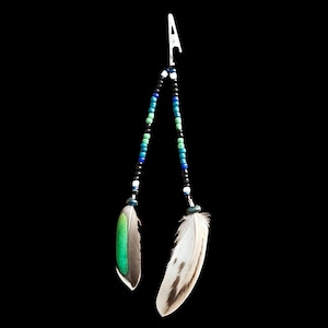 May include: A metal roach clip featuring two dangling strands of beads in shades of blue, green, black, and white. Each strand terminates in a natural feather, with one feather displaying a vibrant green section and the other featuring brown and white markings.