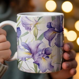May include: A white ceramic mug with a floral design featuring purple and white irises on a white background. The mug has a handle and is being held by a person.