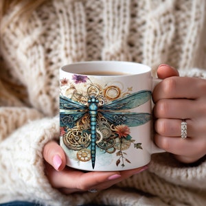 May include: White ceramic mug with a blue and gold dragonfly design. The dragonfly is surrounded by gears and flowers. The mug is filled with coffee.