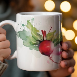 May include: White ceramic mug with a watercolor design of a red beet with green leaves. The mug has a handle and is being held by a hand.