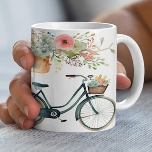 May include: White ceramic mug with a watercolor design of a bicycle with a basket of flowers and a bouquet of flowers spilling out of the basket.