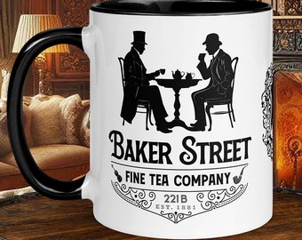 May include: A white ceramic mug with a black rim and handle. The mug features a black and white illustration of two men sitting at a table with a teapot and cups. The text "Baker Street Fine Tea Company 221B Est. 1887" is printed on the mug.
