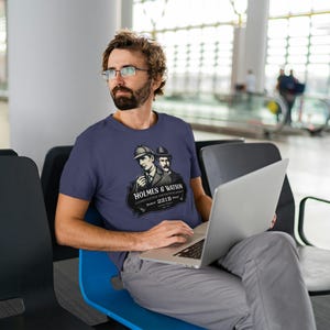 May include: A navy blue t-shirt featuring a graphic of Sherlock Holmes and Dr. Watson with the text "HOLMES & WATSON CONSULTING DETECTIVES Baker Street 221B". The person is sitting and using a laptop.