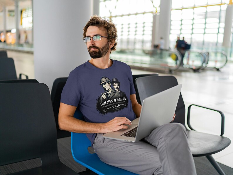 May include: A navy blue t-shirt featuring a graphic of Sherlock Holmes and Dr. Watson with the text "HOLMES & WATSON CONSULTING DETECTIVES Baker Street 221B". The person is sitting and using a laptop.