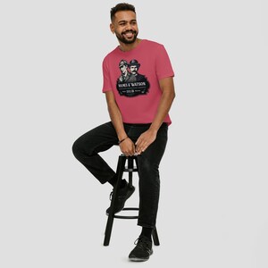May include: A red t-shirt featuring a black and white graphic of two men in hats, with the text "Holmes & Watson" and "221B Baker St." The shirt is being worn by a person sitting on a black stool.