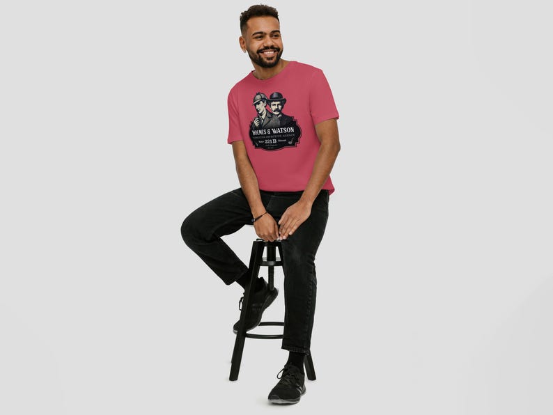 May include: A red t-shirt featuring a black and white graphic of two men in hats, with the text "Holmes & Watson" and "221B Baker St." The shirt is being worn by a person sitting on a black stool.