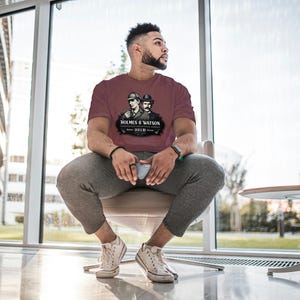 May include: Maroon t-shirt featuring a graphic of Sherlock Holmes and Dr. Watson with the text "HOLMES & WATSON 221B". The shirt is a short-sleeved crew neck.