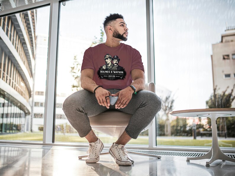 May include: Maroon t-shirt featuring a graphic of Sherlock Holmes and Dr. Watson with the text "HOLMES & WATSON 221B". The shirt is a short-sleeved crew neck.