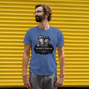 May include: A blue t-shirt featuring a graphic of Sherlock Holmes and Dr. Watson. The graphic includes the text "HOLMES & WATSON" and "CONSULTING DETECTIVE AGENCY" with the address "221B Baker Street". The shirt is worn by a person.