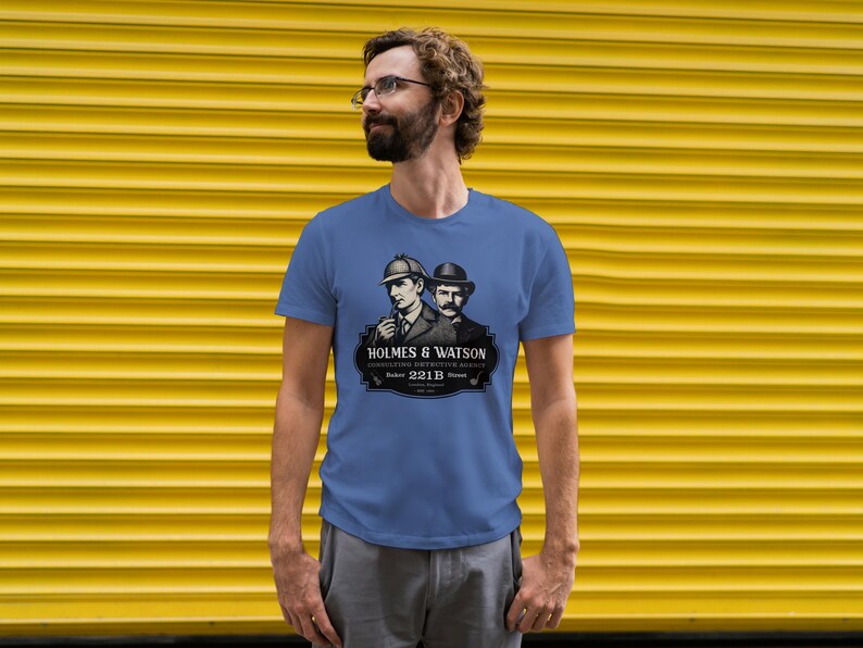 May include: A blue t-shirt featuring a graphic of Sherlock Holmes and Dr. Watson. The graphic includes the text "HOLMES & WATSON" and "CONSULTING DETECTIVE AGENCY" with the address "221B Baker Street". The shirt is worn by a person.