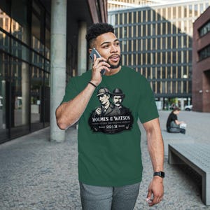May include: Green t-shirt featuring a graphic of Sherlock Holmes and Dr. Watson with the text "HOLMES & WATSON, CONSULTING DETECTIVE AGENCY, 221B Baker Street". The shirt is a short-sleeved crew neck.