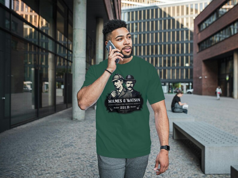 May include: Green t-shirt featuring a graphic of Sherlock Holmes and Dr. Watson with the text "HOLMES & WATSON, CONSULTING DETECTIVE AGENCY, 221B Baker Street". The shirt is a short-sleeved crew neck.