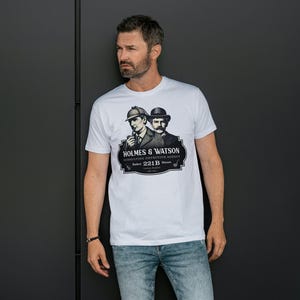 May include: White t-shirt featuring a black and white graphic of Sherlock Holmes and Dr. Watson with the text "HOLMES & WATSON Consulting Detective Agency Baker 221B Street London". The shirt is worn by a person.