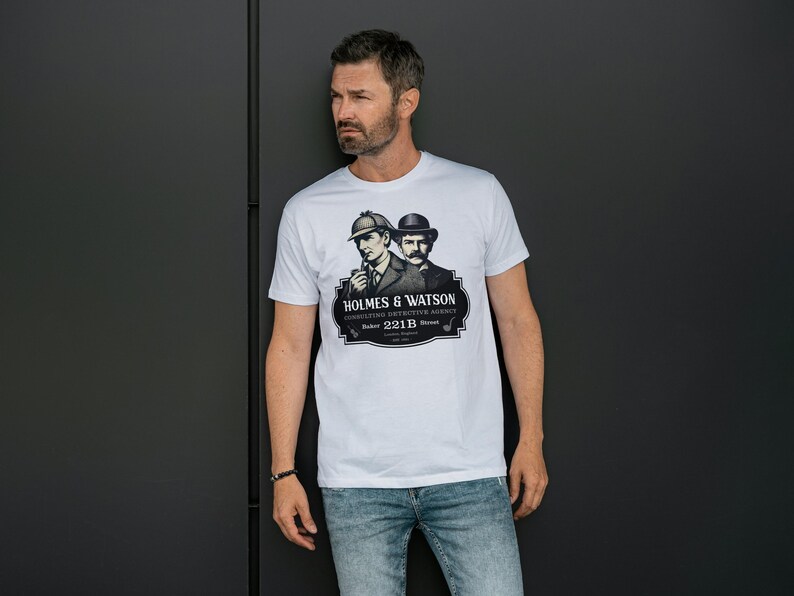 May include: White t-shirt featuring a black and white graphic of Sherlock Holmes and Dr. Watson with the text "HOLMES & WATSON Consulting Detective Agency Baker 221B Street London". The shirt is worn by a person.