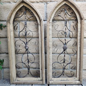May include: Two arched window frames made of light-colored wood, each featuring a central vertical twisted metal rod and decorative black iron scrollwork. The frames have a pointed top and a rectangular base, with a circular metal accent in the center.