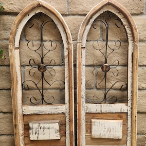 Könnte beinhalten: Zwei rustikale, bogenförmige Wandpaneele aus Holz mit abgenutzter weißer und brauner Farbe. Jedes Paneel hat ein dekoratives schwarzes Metall-Scrollwork-Design und ein kleines, rechteckiges Element. Die Paneele werden vor einer Backsteinmauer ausgestellt.