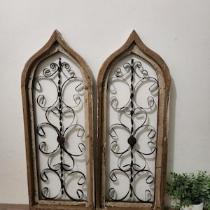 May include: Two decorative wall panels with a distressed wooden frame and a wrought iron design. The ironwork features a symmetrical floral pattern with a central dark brown sphere. The frames are arched at the top, adding a vintage aesthetic.