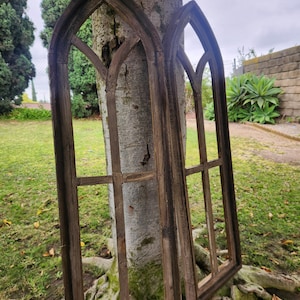 May include: Two tall, dark brown wooden window frames with a pointed gothic arch top. Each frame features a vertical central mullion and a horizontal crossbar, creating a multi-pane structure. The wood has a weathered, textured finish.
