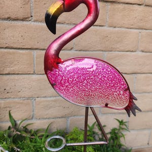 May include: Pink metal flamingo rain gauge with a clear glass tube. The flamingo is standing on a metal stand with a decorative base.
