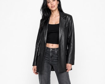 Könnte beinhalten: Ein schwarzer Lederblazer mit steigendem Revers und zwei Vordertaschen mit Patte, getragen über einem schwarzen bauchfreien Top und einer anthrazitfarbenen, gerade geschnittenen Jeans. Das Outfit wird durch schwarze Pumps ergänzt.
