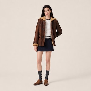 May include: A brown hooded jacket with a tan shearling lining, worn over a white long-sleeve shirt and a dark navy blue mini skirt. The outfit is completed with dark gray crew socks and brown leather loafers, creating a layered look.