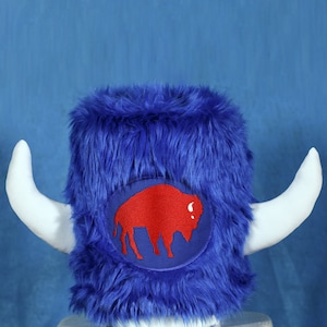 May include: A blue faux fur hat with white horns and a red buffalo logo patch. The hat is sitting on a white stool.