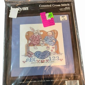 Könnte beinhalten: Gerahmtes Kreuzstich-Set "JUST FRIENDS" von Janlynn. Das Design zeigt ein Kaninchen und ein Baby auf einer Bank, mit einem Herz und den Buchstaben "ABC" und Zahlen "123". Fertige Größe: 25,4 x 25,4 cm.
