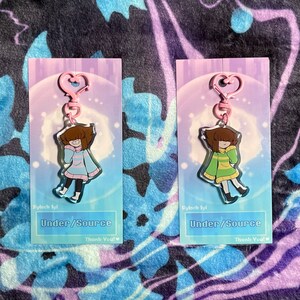 May include: Two pink and white cardstock keychains featuring a cartoon character with brown hair, a blue and white striped sweater, and a green and yellow striped sweater. The text "Stylock fyl" and "Under/Source" is printed on the cards. The text "Thank You!" is printed on the bottom of each card.