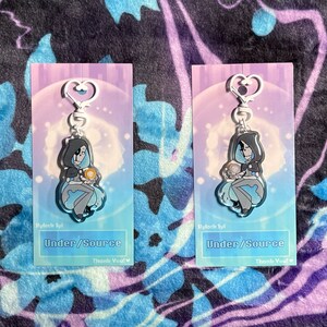 May include: Two clear acrylic keychains featuring a cartoon character with a blue and gray outfit holding a small orb. The keychains are on a pink and blue background with the text "Under/Source" and "Thank You!" printed below the character.
