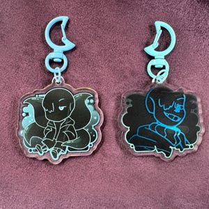 Może przedstawiać: Dwa niebiesko-białe breloki z czarno-białą postacią z kreskówki na każdym. Postać po lewej stronie siedzi, a postać po prawej stronie stoi.