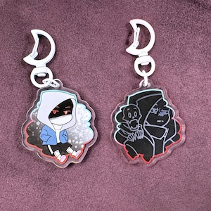 Peut inclure: Deux porte-clés en acrylique représentant des personnages de dessins animés. Le porte-clés de gauche représente un personnage bleu et blanc avec des yeux rouges et une capuche blanche. Le porte-clés de droite représente un personnage noir et blanc avec une capuche noire. Les deux porte-clés ont un anneau métallique blanc.