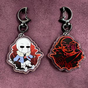May include: Two black and silver keychains with red and black cartoon characters. The keychain on the left features a white character with a blue jacket and red accents. The keychain on the right features a black character with red accents. Both characters have red eyes and are set against a red background.
