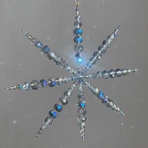 Pode incluir: Um ornamento suspenso em forma de estrela feito de contas de vidro facetadas e iridescentes, transparentes e azul-claras. O ornamento apresenta oito pontas radiantes com contas de vários tamanhos, ligadas por um centro e suspensas por uma fita azul-clara.