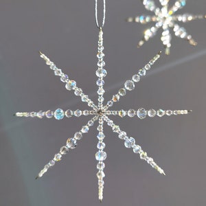 Könnte beinhalten: Zwei klare, sternförmige Ornamente mit schillernden Perlen. Das größere Ornament hat acht Spitzen, jede aus abgestuften Perlen. Zum Aufhängen ist ein dünner, silberner Faden befestigt. Das kleinere Ornament befindet sich im Hintergrund.