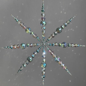 Könnte beinhalten: Ein sternförmiges Ornament aus facettierten, irisierenden Glasperlen in verschiedenen Größen. Das Design besteht aus acht strahlenförmigen Spitzen, die von einem Mittelpunkt ausgehen, mit klaren und bunten reflektierenden Oberflächen.