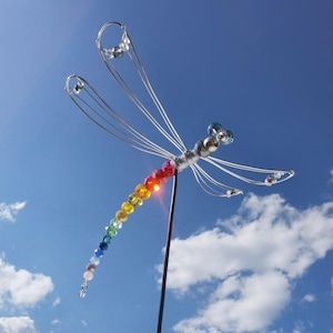 Könnte beinhalten: Eine regenbogenfarbene Perlenlibelle mit Drahtflügeln vor blauem Himmel.