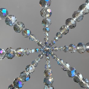 Pode incluir: Um ornamento em forma de estrela composto por contas de cristal transparente facetadas, dispostas em linhas radiantes a partir de uma conta central prateada. As contas t&ecirc;m um acabamento iridescente que reflete a luz com tons azuis e violetas.