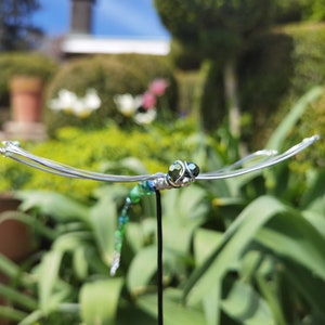 Grüne Deko-Libelle auf Steckstab steckt im Blumenbeet eines Gartens. Ihre Facettenaugen  aus Perlen "blicken" in die Kamera.