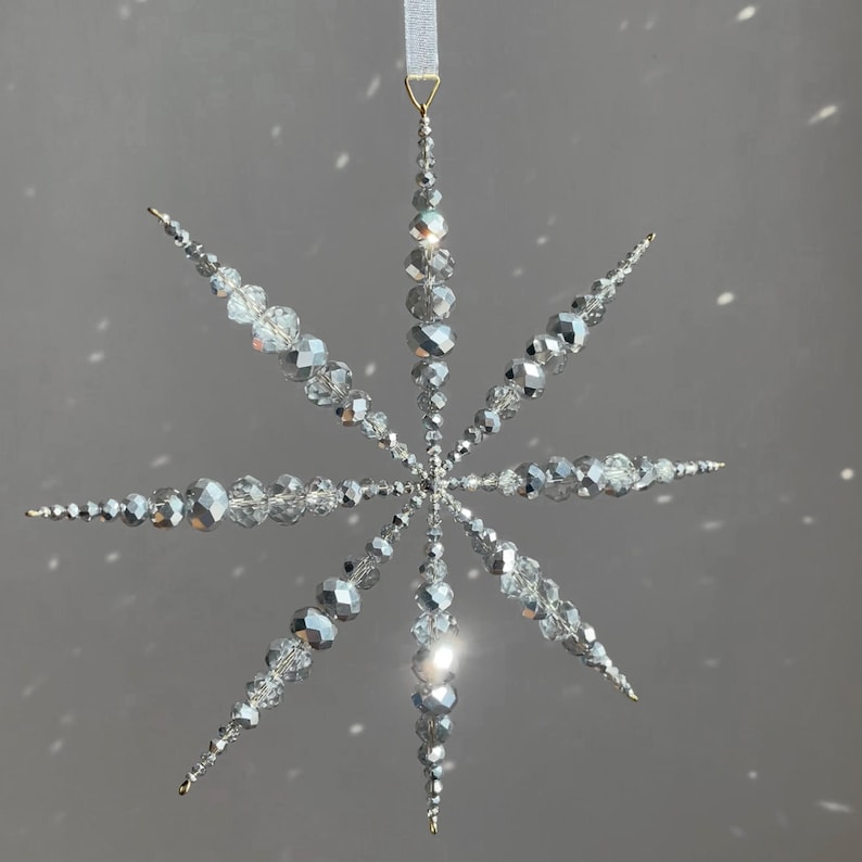 Pode incluir: Um ornamento em forma de estrela feito de contas de vidro facetadas prateadas e transparentes. O design apresenta oito pontas radiantes com contas de v&aacute;rios tamanhos que convergem no centro. Um la&ccedil;o dourado segura uma fita branca.