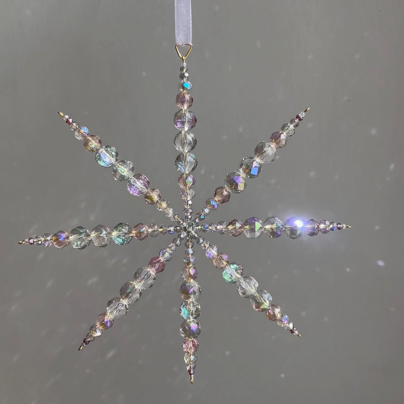 Pode incluir: Um ornamento suspenso em forma de estrela feito de contas de vidro facetado transparentes e iridescentes. O ornamento apresenta oito pontas radiantes com contas de v&aacute;rios tamanhos, ligadas num ponto central e suspensas por uma fita cinzenta clara.