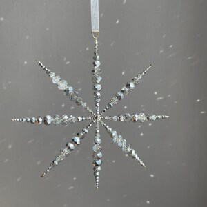Pode incluir: Um ornamento suspenso em forma de estrela composto por contas facetadas prateadas e transparentes dispostas em oito pontas radiantes. O ornamento est&aacute; suspenso por uma fita branca fina presa a um pequeno anel de metal dourado no topo.