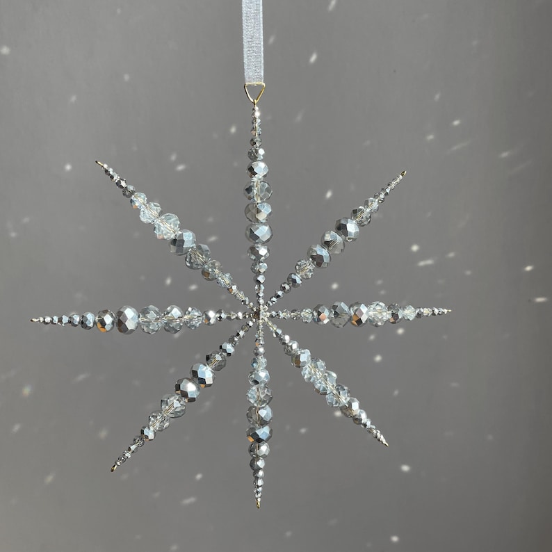 Pode incluir: Um ornamento suspenso em forma de estrela composto por contas facetadas prateadas e transparentes dispostas em oito pontas radiantes. O ornamento est&aacute; suspenso por uma fita branca fina presa a um pequeno anel de metal dourado no topo.